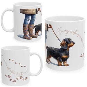 Puede incluir: Taza de cerámica blanca con un perro salchicha con correa y el texto "Cada paseo es una nueva aventura para el alma". La taza también muestra a una persona con vaqueros azules y botas de agua florales. La taza tiene un asa curva.