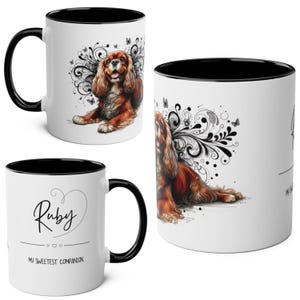 Peut inclure: Mug en céramique blanche avec intérieur et anse noirs. L'illustration détaillée d'un chien Cavalier King Charles Spaniel orne la tasse, avec un motif tourbillonnant décoratif et des papillons. Le mug porte le nom "Ruby" et le texte "My Sweetest Companion".