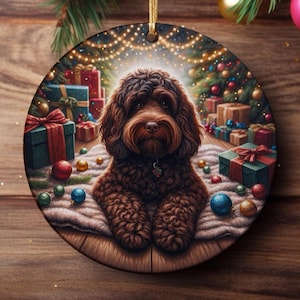 Könnte beinhalten: Ein runder Weihnachtsbaumschmuck aus Keramik mit einem braunen, lockigen Hund, der auf einem weißen Teppich sitzt. Im Hintergrund ist ein geschmückter Weihnachtsbaum mit leuchtenden Lichterketten, bunten Kugeln und mehreren verpackten Geschenken zu sehen.