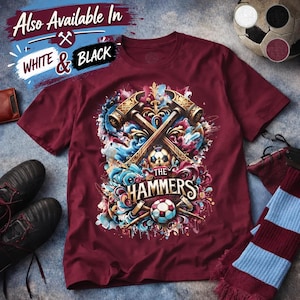 May include: A maroon t-shirt featuring a graphic print of two crossed golden hammers, a soccer ball, and swirling blue and pink abstract shapes. The text "THE HAMMERS" is printed in a bold, stylized font across the center of the design.