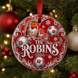 May include: A round red Christmas ornament featuring three robins perched on a branch, three soccer balls, and white snowflake accents. The text reads THE ROBINS 1894 in a decorative font. The ornament hangs from a red ribbon loop.