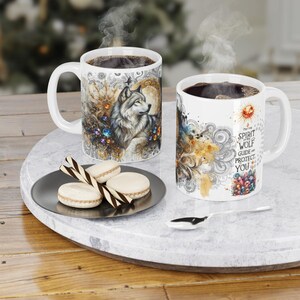 May include: Two white ceramic mugs with coffee, featuring wolf and floral designs. One mug has the text "The Spirit Wolf Guide and Protect You". A plate with macarons and a spoon are on a marble tray.