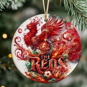 May include: A round ceramic ornament featuring a vibrant red phoenix illustration, a gold-toned Christmas bauble, and a soccer ball graphic. The text THE REDS is printed in bold red lettering amidst swirling flames and abstract red and green patterns.