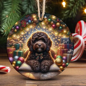 May include: A round Christmas ornament featuring a fluffy brown dog wearing a plaid bow tie, resting on a pillow. The scene includes colourful wrapped gift boxes, shiny ornaments, and glowing string lights against a festive evergreen background.
