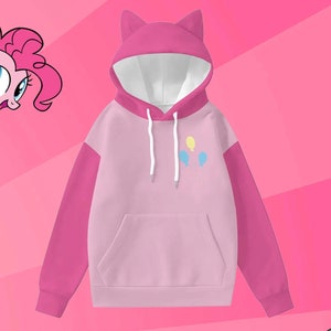 Puede incluir: Sudadera con capucha rosa y rosa claro con orejas de gato en la capucha. Las mangas son de un rosa más oscuro. La parte delantera de la sudadera tiene un bolsillo de canguro y un pequeño gráfico de tres globos. Un personaje de dibujos animados está en la esquina superior izquierda.