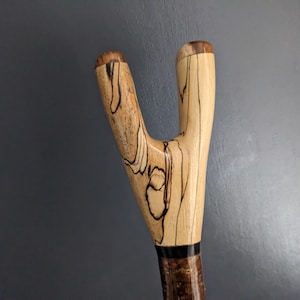 May include: A wooden walking stick handle featuring a Y-shaped design with a smooth, polished finish. The wood displays natural light tan tones with dark, intricate grain patterns and black streaks. A thin black band separates the handle from the shaft.