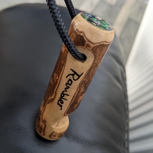 May include: A cylindrical wooden pendant with a natural grain pattern, featuring the word Rambler engraved in black script. A black cord passes through a hole near the top, and a small circular compass with a green and silver face is embedded.