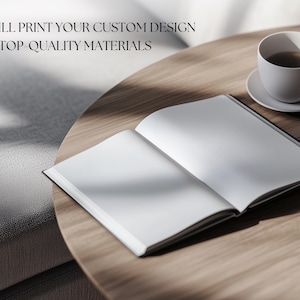 May include: A blank, white book with a black spine is open on a wooden table. A white coffee cup with a brown liquid inside is on a white saucer to the right of the book. The text "WE WILL PRINT YOUR CUSTOM DESIGN WITH TOP-QUALITY MATERIALS" is visible at the top of the image.