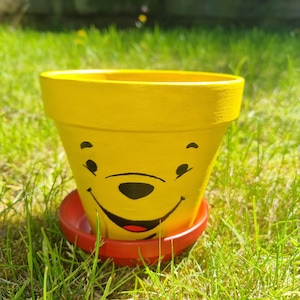 May include: A yellow painted flower pot with a smiling face design, sitting on a red saucer. The pot is outdoors on a bed of green grass.