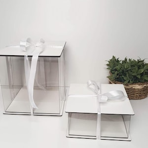 Könnte beinhalten: Zwei transparente Acryl-Geschenkboxen mit weißen Deckeln und weißem Band. Die größere Box ist 30 cm hoch und die kleinere 20 cm hoch.