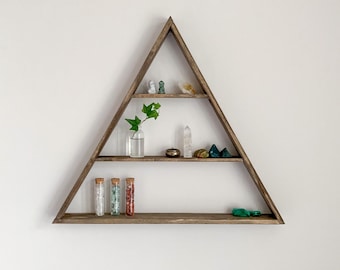 Puede incluir: Una estantería de madera triangular con tres estantes. El estante superior tiene una pequeña figurita blanca, una figurita verde y algunos cristales pequeños. El estante del medio tiene un pequeño jarrón de vidrio con una planta verde, una pequeña caja de madera y algunos cristales. El estante inferior tiene tres pequeños tubos de vidrio llenos de cristales. La estantería está contra una pared blanca.