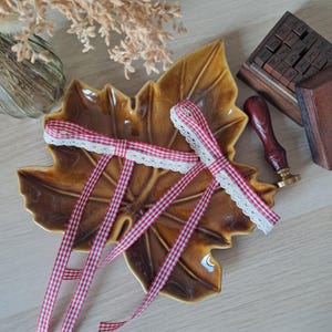 Peut inclure: Deux rubans à carreaux rouges et blancs avec une bordure en dentelle blanche reposent sur un plat en céramique en forme de feuille marron. Un jeu de tampons en bois et un tampon à cacheter à manche rouge sont également visibles.