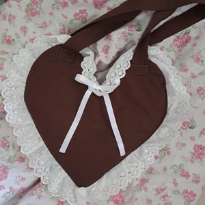 Peut inclure: Un sac à main marron en forme de cœur avec une bordure en dentelle blanche et un nœud en ruban blanc. Le sac a des bretelles marron et est posé sur un fond floral. La garniture en dentelle ajoute une touche délicate au design.