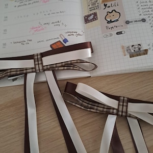 以下が含まれることがあります： 茶色、白、チェック柄のリボンが付いた2つの装飾的なリボン。リボンは、手書きのメモとステッカーが付いたノートに配置されています。ノートには、日付16が表示されたカレンダーレイアウトがあります。