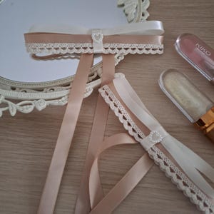 以下が含まれることがあります： アイボリーとブラッシュサテンのリボンを使用した装飾的なヘアアクセサリーのクローズアップ。繊細なレースのトリムと小さなハート型の装飾が施されています。アクセサリーは木製の表面に配置され、背景には鏡とリップグロスが見えます。