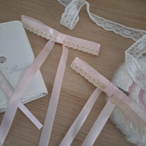 以下が含まれることがあります： ピンクのサテンリボン、クリーム色のレーストリム、白い合皮ケースなどのクラフト用品のコレクション。リボンはクリーム色のレーストリム付きで、リボン結びになっています。明るい木製の表面に配置されています。