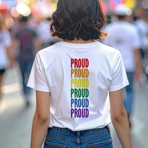 May include: A white t-shirt with the word "PROUD" in rainbow colors down the back. The shirt is worn by a person in a crowd, with rainbow flags and other people in the background. The image captures a moment of pride and celebration.