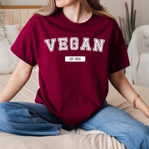 Peut inclure: Un t-shirt bordeaux avec le mot "VEGAN" en blanc, en lettres de style universitaire. En dessous du mot "VEGAN" se trouve le texte "EST. 2024" dans un rectangle blanc. Le t-shirt est à manches courtes.