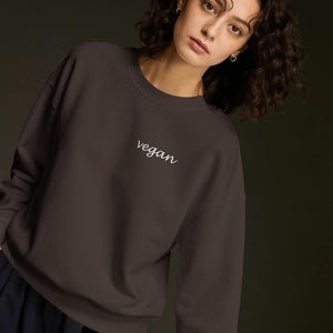 Op de afbeelding: Een donker antracietgrijze sweater met ronde hals en het woord vegan in witte cursieve letters geborduurd in het midden van de borst. Het kledingstuk heeft lange mouwen en een geribbelde halslijn voor een eenvoudige uitstraling.