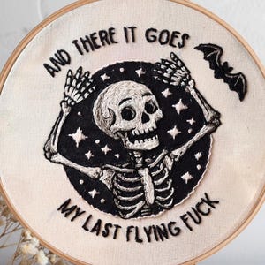 Könnte beinhalten: Gesticktes Stickrahmen-Kunstwerk mit einem Skelett mit erhobenen Armen, umgeben von einem schwarzen Kreis mit Sternen. Der Text "AND THERE IT GOES MY LAST FLYING FUCK" ist oberhalb und unterhalb des Skeletts gestickt. Eine kleine Fledermaus ist rechts gestickt.