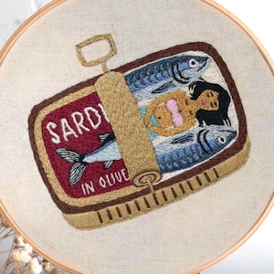 May include: Embroidered artwork in a wooden hoop. The design depicts a sardine can with the word "SARDI" and "IN OLIVE" on the label. Inside the can are fish and a mermaid. The can is brown and red, with blue fish and a mermaid in a pink bikini.