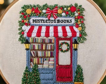 Könnte beinhalten: Gesticktes Hoop-Art mit einer skurrilen Buchladen-Szene. Der Laden hat eine rot-weiß gestreifte Markise, eine rote Tür und Regale voller Bücher. Das Schild darüber lautet "MISTLETOE BOOKS". Das Design ist von festlichem Grün und einer roten Schleife umgeben.