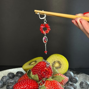 Könnte beinhalten: Hängende Ohrringe mit roten Edelsteinakzenten, die von Essstäbchen gehalten werden. Die Ohrringe befinden sich über einem Teller mit frischen Erdbeeren, Blaubeeren und Kiwischeiben. Der Hintergrund ist dunkelgrau.