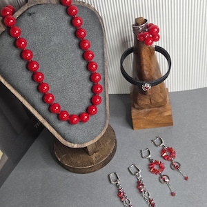Könnte beinhalten: Ein Schmuckset bestehend aus einer Kette aus runden roten Perlen, einem schwarzen Lederarmband mit rotem Perlenakzent, einem Ring mit rotem Perlencluster sowie zwei Paar Ohrhängern mit roten Kristallperlen und silberfarbenen Metallketten.