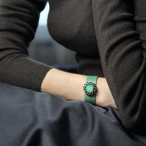 Könnte beinhalten: Ein grünes Lederarmband mit einem kreisförmigen, bronzefarbenen Verschluss. Das Armband wird am Handgelenk einer Person getragen, mit einem dunkelbraunen Langarmshirt und einem dunkelgrauen Rock im Hintergrund.