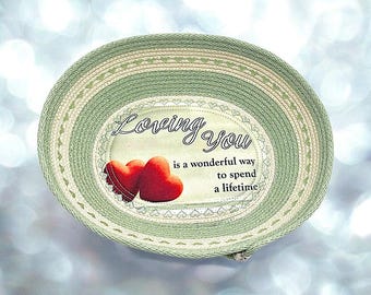 May include: A green woven bowl with a white and green striped design. The bowl has a heart-shaped design on the rim and a fabric insert with the text "Loving You is a wonderful way to spend a lifetime" and two red hearts.