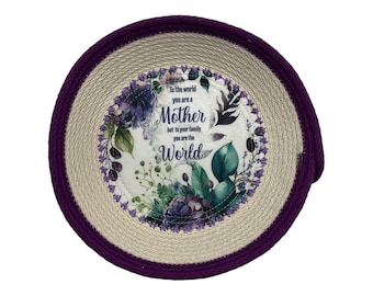 May include: A round, woven, purple and beige coaster with a white centre featuring a floral design and the text "To the world you are a Mother but to your family you are the World."