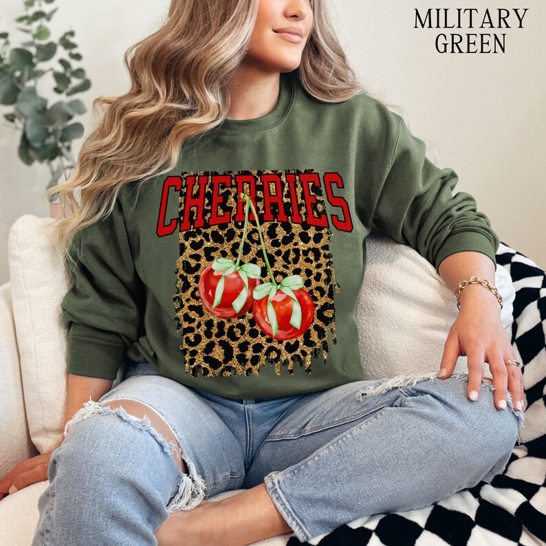 May include: A green sweatshirt with the word "CHERRIES" printed in red on the front. The word is surrounded by a leopard print design with two red cherries with green stems.