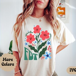 Puede incluir: Una camiseta blanca con un diseño floral y el texto "FDT" en verde. Las flores son rojas, azules y verdes. La camiseta está disponible en marfil.
