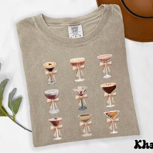 Pode incluir: Camiseta c&aacute;qui com ilustra&ccedil;&otilde;es em aquarela de coquet&eacute;is, cada um com um la&ccedil;o. A camiseta tem gola redonda e &eacute; feita de um material macio. A palavra "Khaki" est&aacute; impressa na parte inferior. A camiseta tem um ajuste confort&aacute;vel.