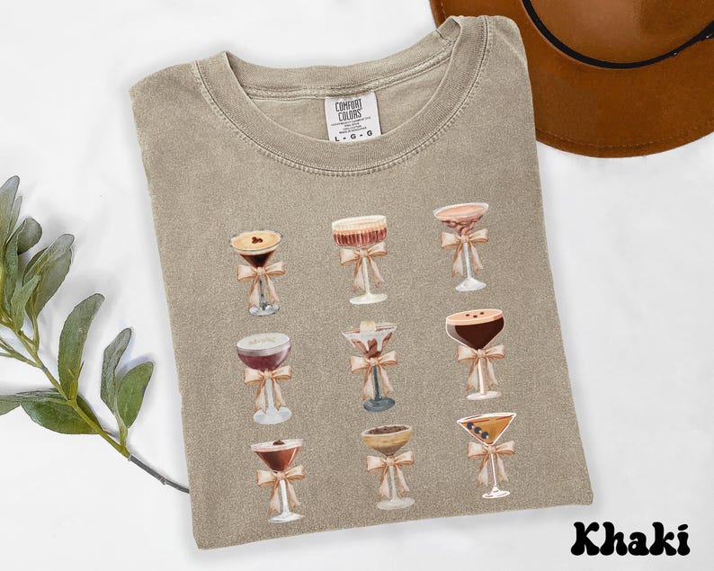 Pode incluir: Camiseta c&aacute;qui com ilustra&ccedil;&otilde;es em aquarela de coquet&eacute;is, cada um com um la&ccedil;o. A camiseta tem gola redonda e &eacute; feita de um material macio. A palavra "Khaki" est&aacute; impressa na parte inferior. A camiseta tem um ajuste confort&aacute;vel.