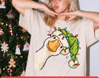 Pode incluir: Uma t-shirt cor de marfim com uma ilustração gráfica de um ganso branco a usar um gorro de Pai Natal verde adornado com luzes de Natal coloridas. O ganso tem um bico cor de laranja e uma borda branca felpuda no gorro.