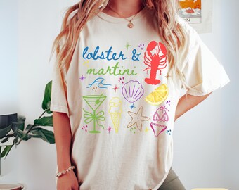 May include: Ivory-colored t-shirt with a colorful graphic design. The design includes the text "lobster & martini" along with illustrations of a lobster, martini glass, seashell, ice cream cone, starfish, lemon slice, and bikini.