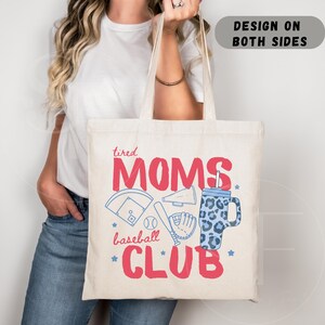Op de afbeelding: Crèmekleurige draagtas met de tekst "tired MOMS baseball CLUB" in rood en blauw. Het ontwerp bevat honkbalafbeeldingen en een blauwe beker met luipaardprint. De tekst "DESIGN ON BOTH SIDES" staat bovenaan.