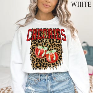 May include: White sweatshirt with the word "CHERRIES" printed in red and black letters. The word is on top of a leopard print design with gold glitter. Two red cherries with green stems and leaves are on the leopard print.
