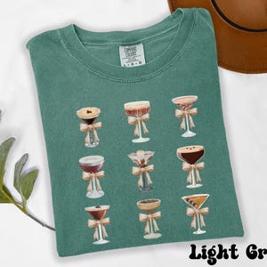 Pode incluir: Camiseta verde claro com um design em aquarela de coquet&eacute;is, cada um adornado com um la&ccedil;o. A camisa &eacute; rotulada como "Comfort Colors" e o texto "Light Green" est&aacute; na parte inferior. A camisa est&aacute; dobrada em uma superf&iacute;cie branca.