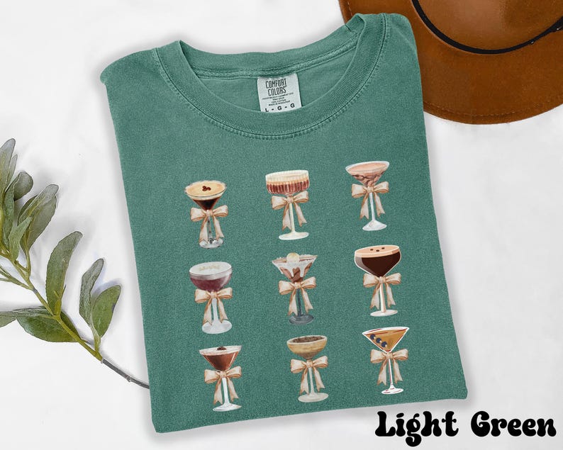 Pode incluir: Camiseta verde claro com um design em aquarela de coquet&eacute;is, cada um adornado com um la&ccedil;o. A camisa &eacute; rotulada como "Comfort Colors" e o texto "Light Green" est&aacute; na parte inferior. A camisa est&aacute; dobrada em uma superf&iacute;cie branca.