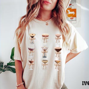 Pode incluir: Camiseta de cor marfim com ilustra&ccedil;&otilde;es de coquet&eacute;is em copos transparentes, cada um adornado com um la&ccedil;o. O design apresenta uma variedade de coquet&eacute;is, com diferentes cores e texturas. A palavra "IVORY" est&aacute; impressa em negrito. A camiseta &eacute; de cor neutra.