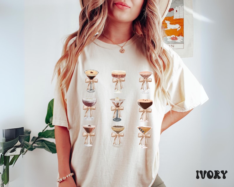 Pode incluir: Camiseta de cor marfim com ilustra&ccedil;&otilde;es de coquet&eacute;is em copos transparentes, cada um adornado com um la&ccedil;o. O design apresenta uma variedade de coquet&eacute;is, com diferentes cores e texturas. A palavra "IVORY" est&aacute; impressa em negrito. A camiseta &eacute; de cor neutra.