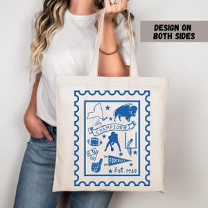 Può includere: Borsa tote color crema con un design blu a forma di timbro. Il design presenta un tema calcistico con un bufalo, un pallone da football e il testo "CHAMPION #1 FOOTBALL Est. 1960". Le parole "DESIGN ON BOTH SIDES" sono stampate in alto a destra.