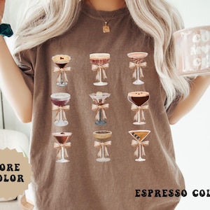 Pode incluir: Camiseta cor caf&eacute; com um desenho em aquarela de nove ta&ccedil;as de coquetel, cada uma adornada com um la&ccedil;o. O texto "MORE COLOR" est&aacute; em uma estrela bege, e "ESPRESSO COLOR" est&aacute; na parte inferior.