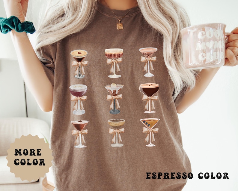 Pode incluir: Camiseta cor caf&eacute; com um desenho em aquarela de nove ta&ccedil;as de coquetel, cada uma adornada com um la&ccedil;o. O texto "MORE COLOR" est&aacute; em uma estrela bege, e "ESPRESSO COLOR" est&aacute; na parte inferior.