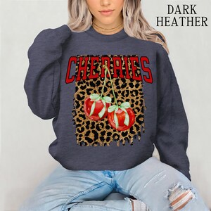 May include: Dark heather grey sweatshirt with a red "Cherries" graphic on a gold glitter leopard print background. The graphic features two red cherries with green stems and leaves.