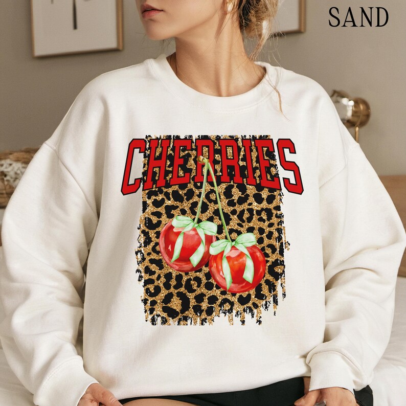 May include: White sweatshirt with a graphic print of two red cherries on a gold glitter leopard print background. The text "CHERIES" is printed in red above the cherries.