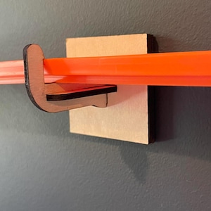 May include: A wooden track holder mounted on a gray wall, designed to hold a bright orange toy car track. The holder is made of light brown wood and has a simple, functional design. The track is approximately 1 inch wide.