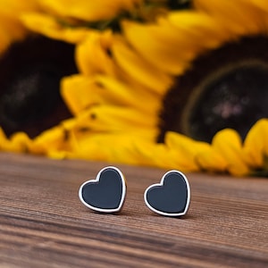 Puede incluir: Un par de pendientes de corazón negro con engastes plateados. Los pendientes están sobre una superficie de madera con un fondo borroso de girasoles amarillos.